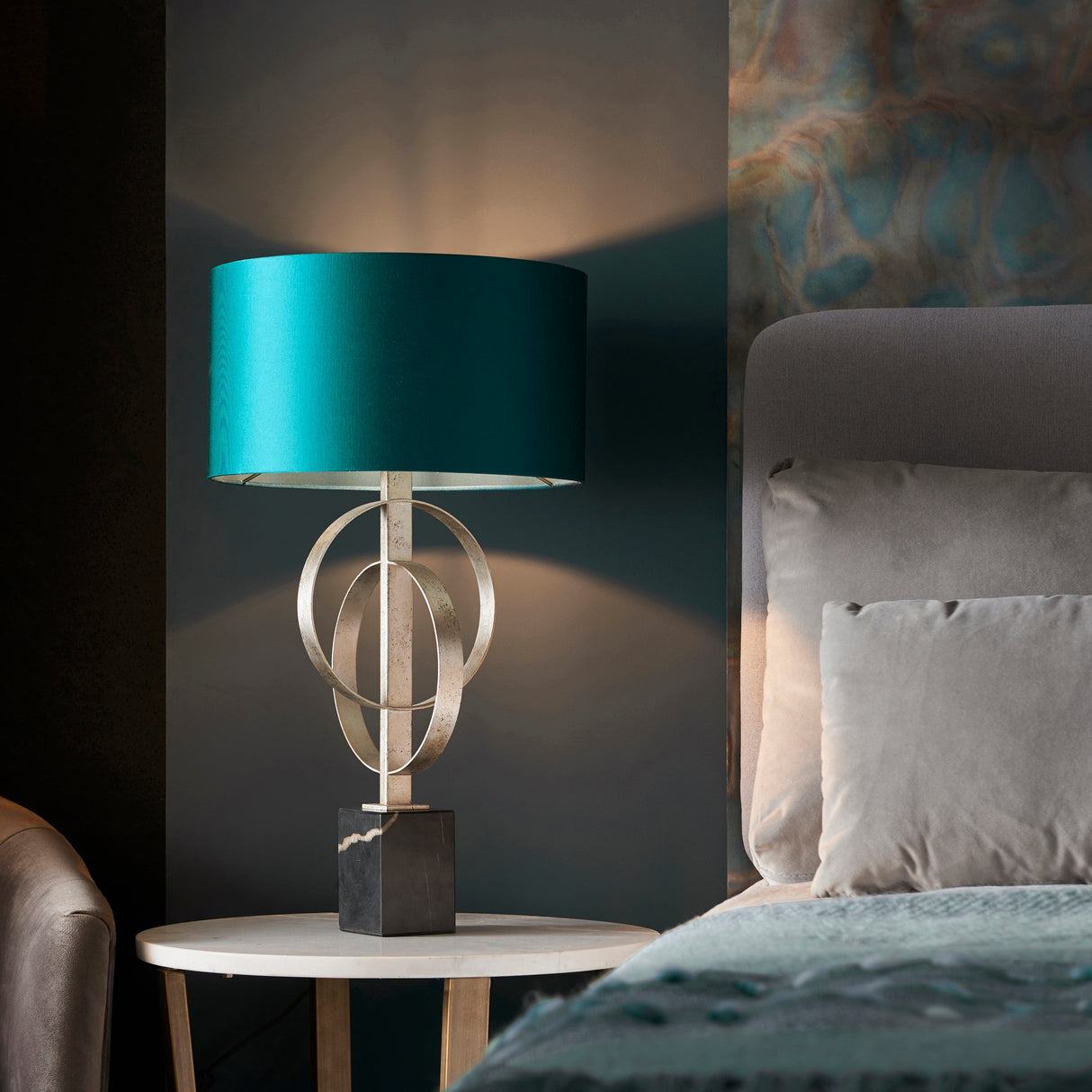 Sparrowmere Double Hoop table lamp in silver leaf with teal drum shade and marble base, on a bedside table beside a grey upholstered bed.