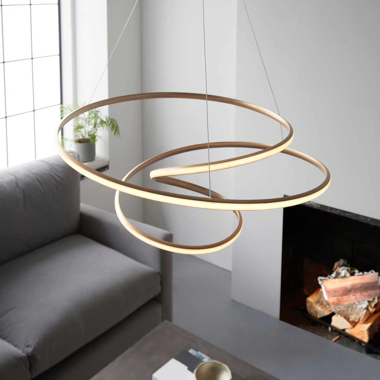 Tolmere Spiral Large Pendant in natural metal finish, coiled ribbon LED fitting suspended above a grey sofa beside a fireplace in a living room.