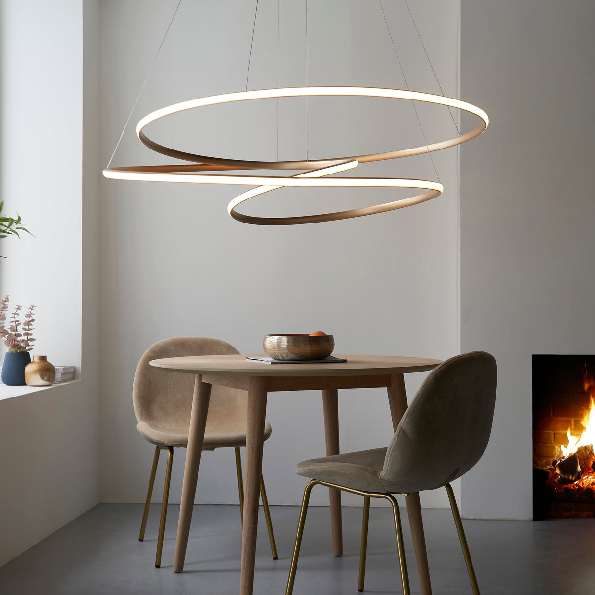 Tolmere Spiral Grand Pendant in natural metal finish by Lighting No.1, illuminated above a round wooden dining table with velvet chairs beside a fireplace.