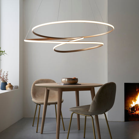 Tolmere Spiral Grand Pendant in natural metal finish by Lighting No.1, illuminated above a round wooden dining table with velvet chairs beside a fireplace.
