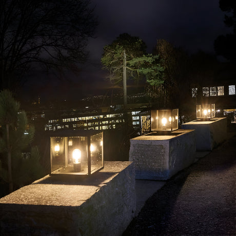 A stone pathway is illuminated by the Lofoten 1 Light Pedestal - Black, cube-shaped lanterns casting a glow against the trees and dark night sky. This contemporary outdoor lighting creates a warm, inviting atmosphere, perfectly contrasting with the surrounding shadows in sophisticated outdoor spaces.