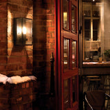 The warmly lit entrance with a wooden door slightly open reveals a cozy café, its rustic charm enhanced by brick walls and snow on the exterior ledge. The Lofoten Outdoor 1 Light Half Lantern in black, featuring an IP44 rating, casts a glow inside where you can see a bar counter, stools, and a potted plant through the window.