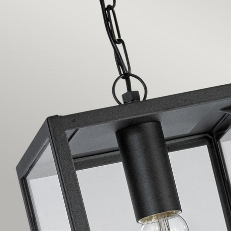 A close-up view of the Lofoten Outdoor 1 Light Chain Pendant in black showcases its industrial elegance. This pendant light features a modern, rectangular design with clear glass panels revealing a visible bulb inside. It is suspended by a black metal chain against a gray background and boasts an IP44 rating for durability.