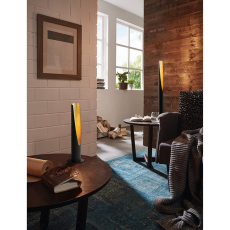 Losessa Floor Lamp in black and gold, slim steel tube with angled cut-out revealing warm gold glow, shown lit in a living room beside a table and armchair.