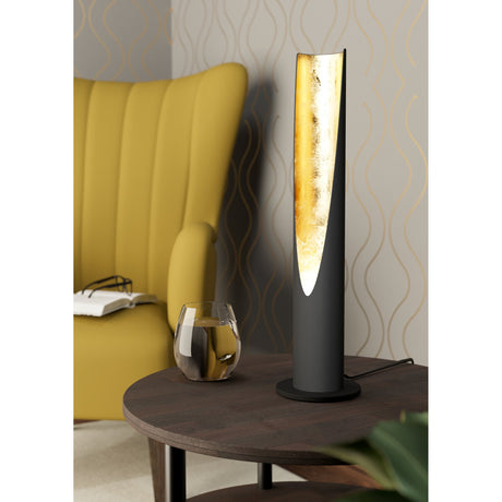 Losessa Table Lamp in black steel with a tall angled cut-out revealing a gold-leaf interior, shown lit on a round base beside a yellow armchair.