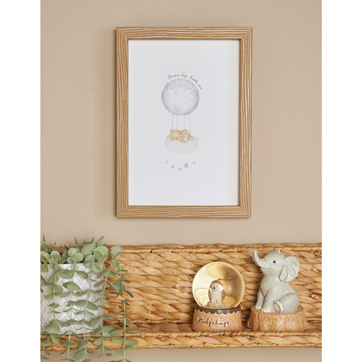 Framed A4 wall art in a natural wood frame, shown on a beige wall above a woven shelf, depicting a watercolour moon balloon lifting a sleeping bear on a cloud with stars.