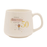 White stoneware birthday mug with gold "50" and "Blooming Beautiful" text, pink brushstroke accent, and an illustrated flower-holding figure with hearts, shown on a plain white background.