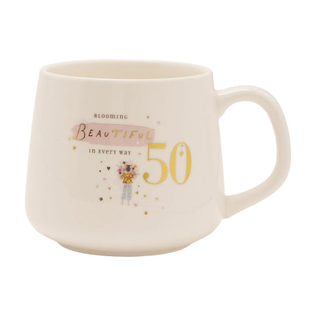 White stoneware birthday mug with gold "50" and "Blooming Beautiful" text, pink brushstroke accent, and an illustrated flower-holding figure with hearts, shown on a plain white background.