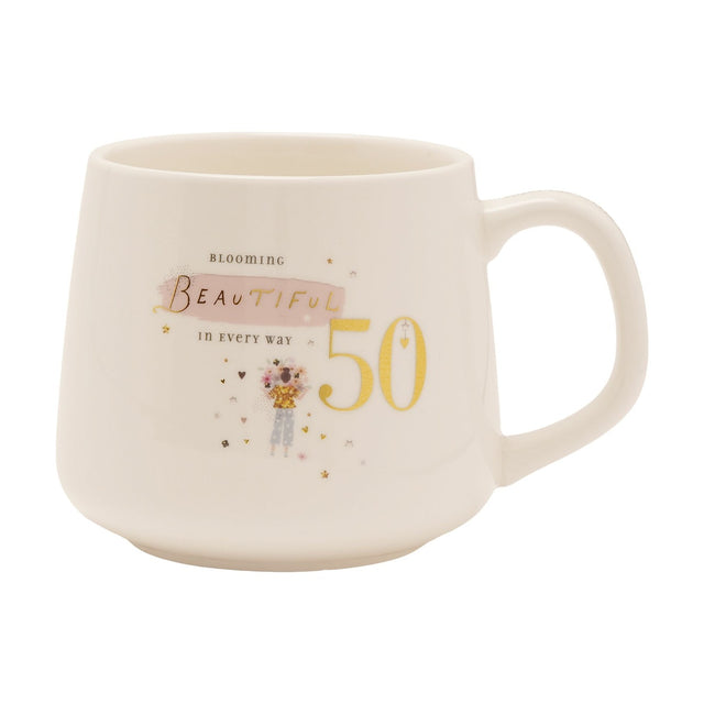White stoneware birthday mug with gold "50" and "Blooming Beautiful" text, pink brushstroke accent, and an illustrated flower-holding figure with hearts, shown on a plain white background.
