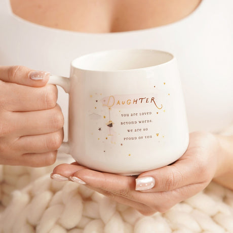 White stoneware mug held in hands, printed with "Daughter, you are loved beyond words" in gold script with a swing figure, cloud, and star details.