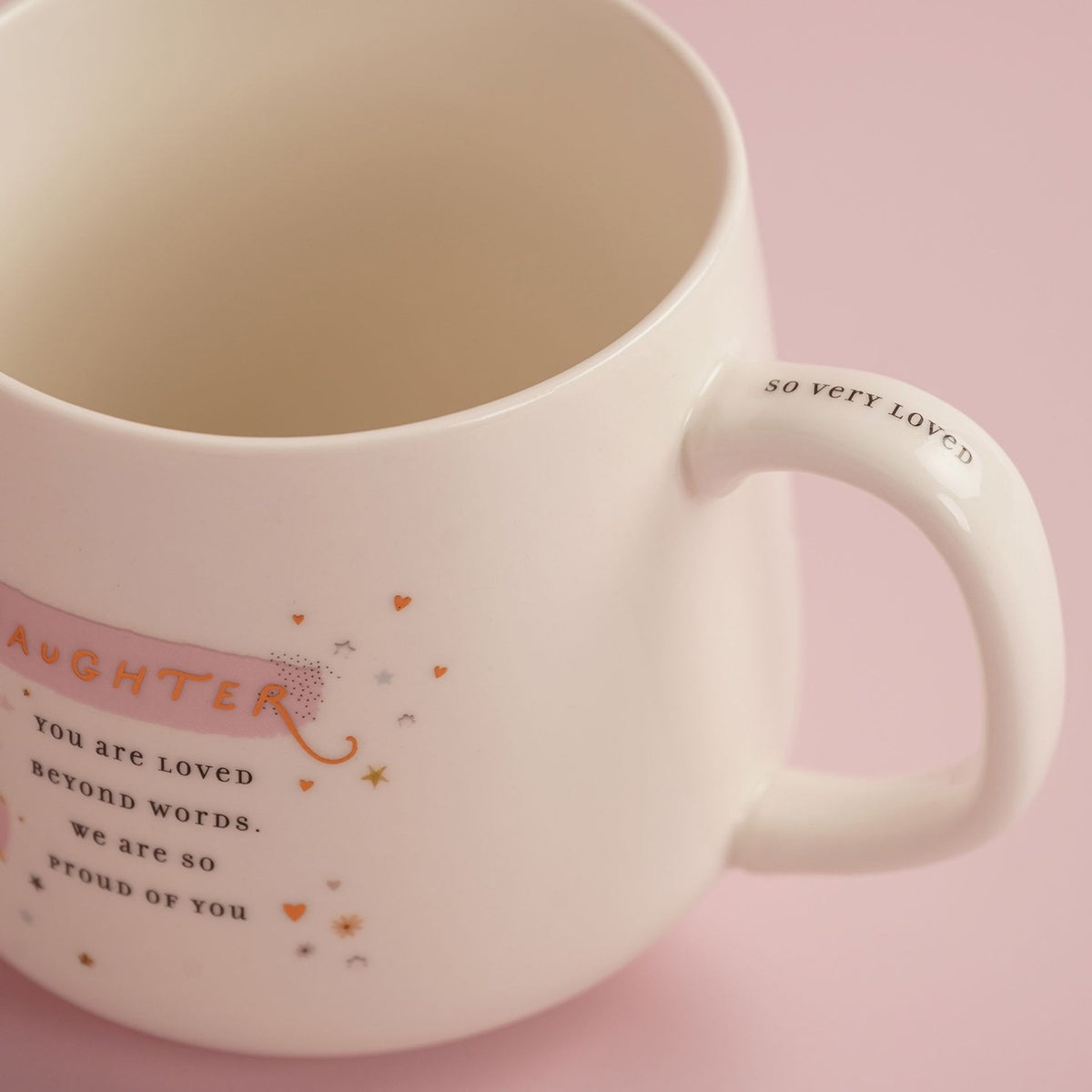 White stoneware mug with pink "Daughter" banner, gold script text "You are loved beyond words, we are so proud of you," stars, hearts and "So very loved" printed on the handle.