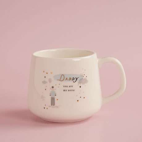 Cream stoneware mug with C-handle, printed Daddy you are my hero in gold script with dad carrying child, balloon, clouds and gold hearts, on pink background.