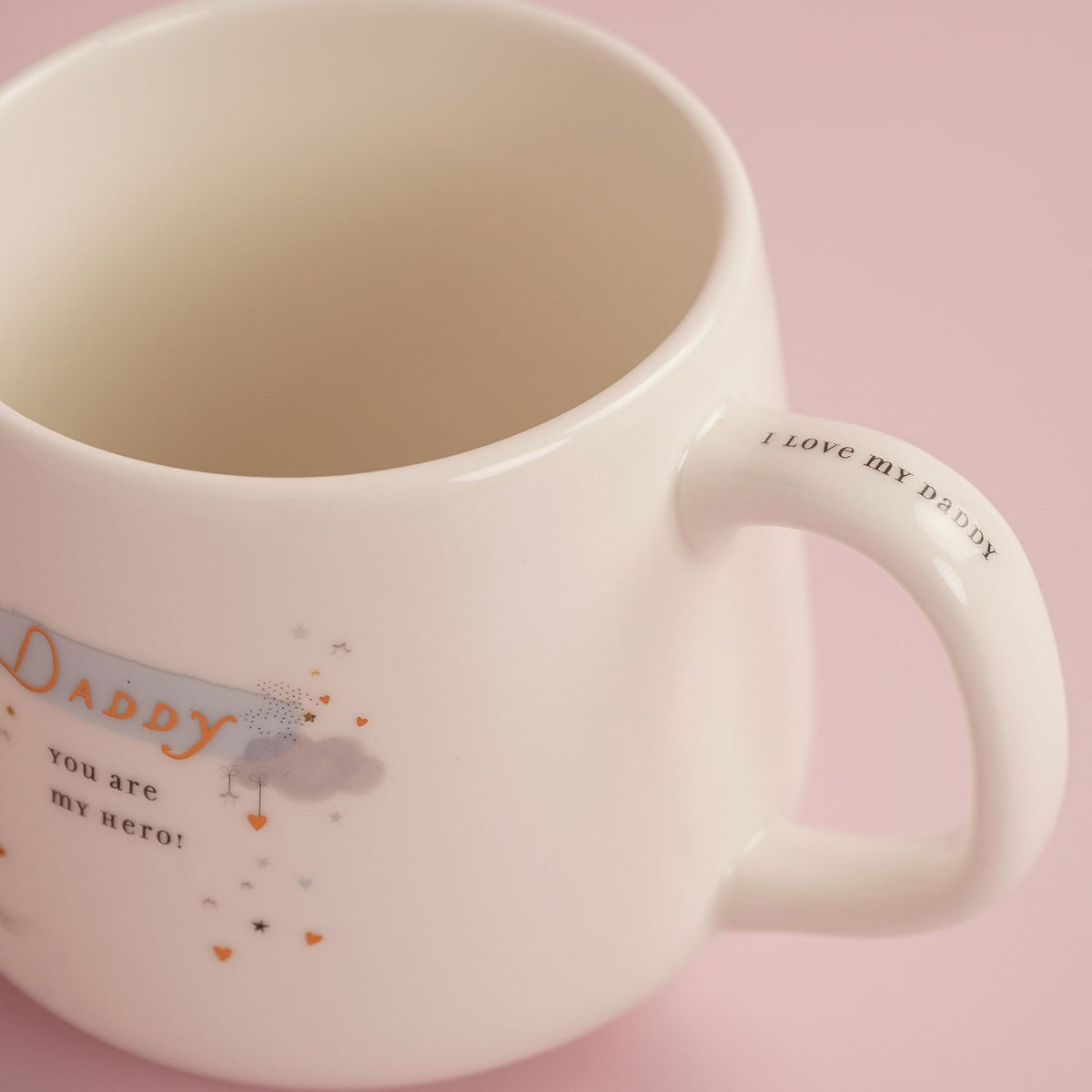 Close-up of a cream stoneware mug with a curved handle printed "I love my Daddy", and "Daddy you are my hero" text with hearts and stars on the body, set against a pink background.