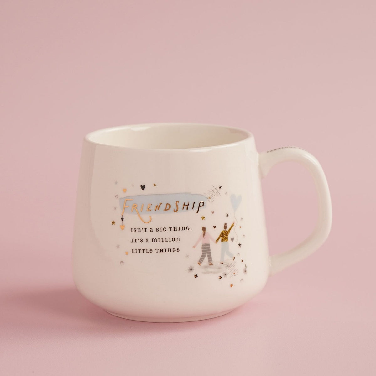 White stoneware mug with handle, featuring "Friendship" in gold lettering over a blue brushstroke, two figures with a heart balloon, hearts and stars, on a pink background.