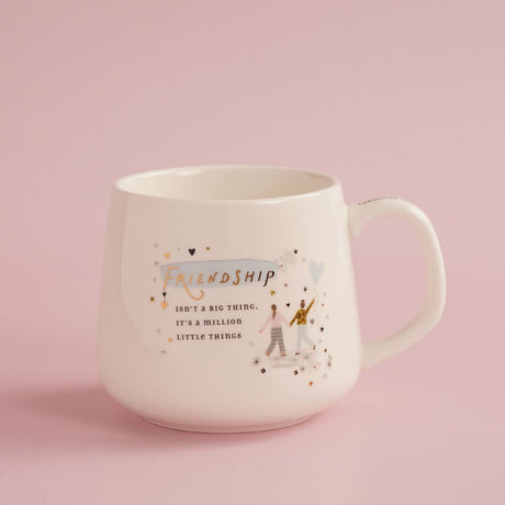 White stoneware mug with handle, featuring "Friendship" in gold lettering over a blue brushstroke, two figures with a heart balloon, hearts and stars, on a pink background.