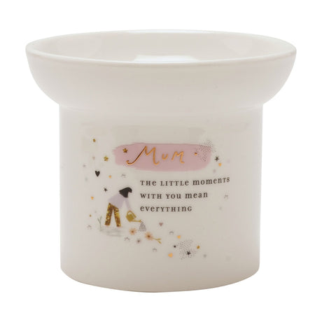 White round ceramic wax melt burner with gold "Mum" lettering on pink brushstroke, a woman watering flowers illustration, gold stars and hearts, on a plain background.