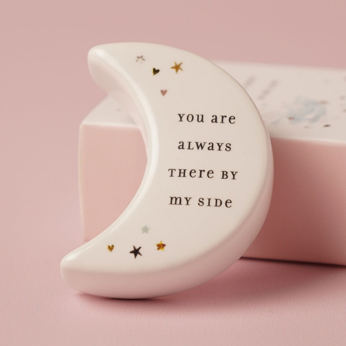 White ceramic crescent moon token from the Auntie Moon Keepsake Box, reverse side reading "You are always there by my side" with small star and heart accents, on a pink background.
