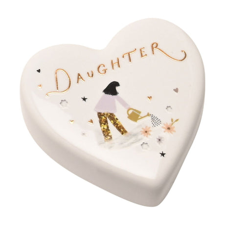 White ceramic heart-shaped keepsake box with "Daughter" in gold lettering, featuring an illustrated girl with dark hair watering pink flowers with a gold glitter accent, on a plain white background.