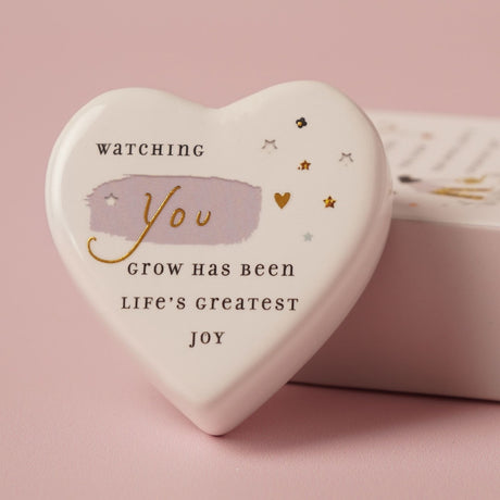 White ceramic heart-shaped keepsake token with matchbox, printed with "Watching You grow has been life's greatest joy" in black and gold text, star and heart details, on a pink background.