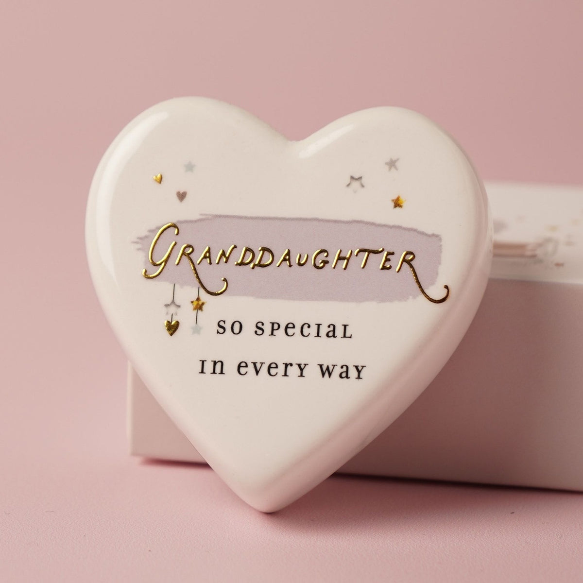 White ceramic heart keepsake reading Granddaughter, so special in every way, with gold stars and hearts over a lilac brushstroke, shown with its matchbox on a pink background.
