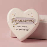 White ceramic heart keepsake reading Granddaughter, so special in every way, with gold stars and hearts over a lilac brushstroke, shown with its matchbox on a pink background.