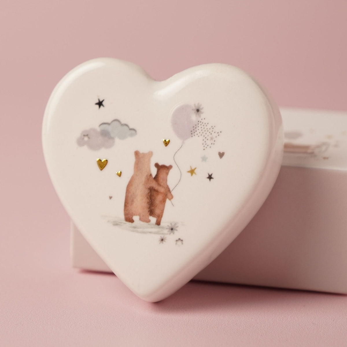 White ceramic heart-shaped keepsake token with two bears holding a balloon, surrounded by stars and hearts, resting on its matchbox against a pink background.