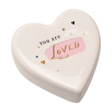 White ceramic heart-shaped keepsake token reading "You are LOVED" in black and gold lettering over a pink brushstroke, with small gold hearts and stars, on a plain white background.