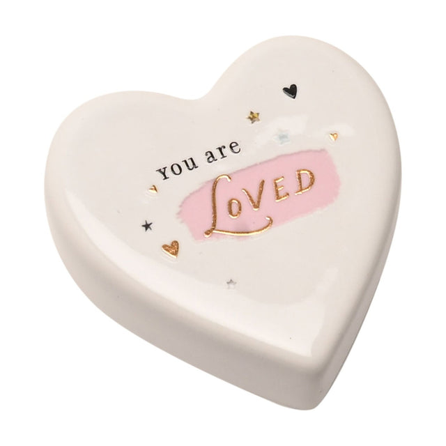 White ceramic heart-shaped keepsake token reading "You are LOVED" in black and gold lettering over a pink brushstroke, with small gold hearts and stars, on a plain white background.