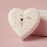 White ceramic heart keepsake box showing the back illustration of a girl on a swing with stars, clouds, and gold hearts, set against a pink background.