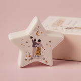 White ceramic star-shaped keepsake token with an illustration of two figures embracing under a crescent moon, surrounded by hearts and flowers, shown beside its matching box on a pink background.
