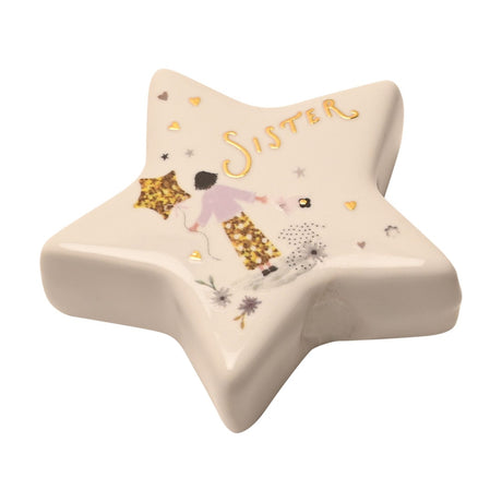White ceramic star-shaped keepsake token with glossy finish, featuring "Sister" in gold script above an illustrated girl in a lilac top holding a star balloon, surrounded by small hearts and flowers.