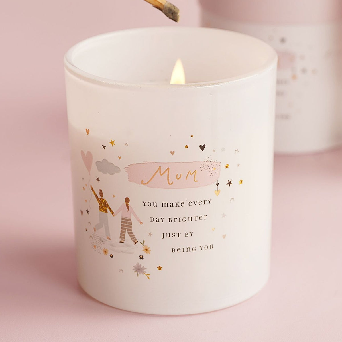 Lit round white jar scented candle for Mum with pink brushstroke, gold "Mum" lettering, hand-in-hand figures with heart balloon, and scattered gold stars.