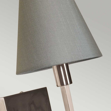 Close-up of the Lucerne 1 Light Wall Light featuring a brushed nickel finish on its silver metallic base and a gray conical fabric shade. The light gray background complements the lamp's contemporary vibe and minimalist design.