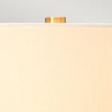 Close-up of a beige lampshade against a light gray background. Visible above its recycled glass base, the top of the Luga Table Lamp - Aged Brass showcases elegant aged brass detailing, highlighting its eco-conscious design.