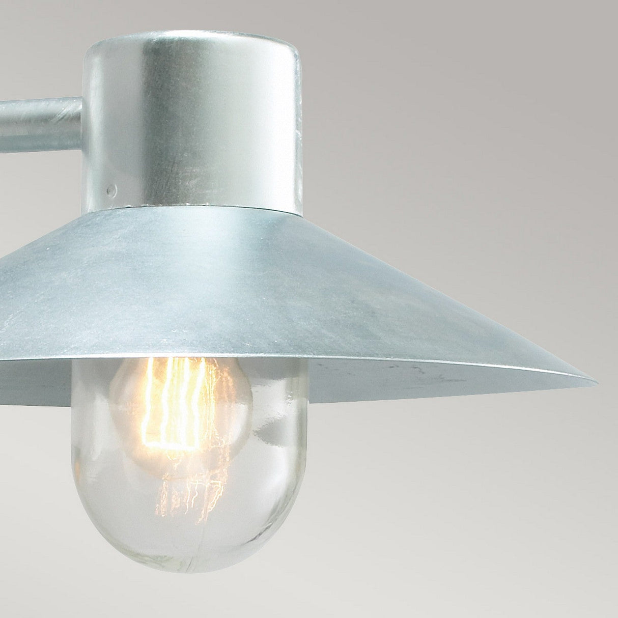 Close-up of the Lund Outdoor 1 Light Wall Lantern in a sleek silver design. The exposed bulb emits a warm glow, protected by a conical shade. Made from galvanized steel, this IP65 rated fixture is beautifully set against a neutral grey backdrop.