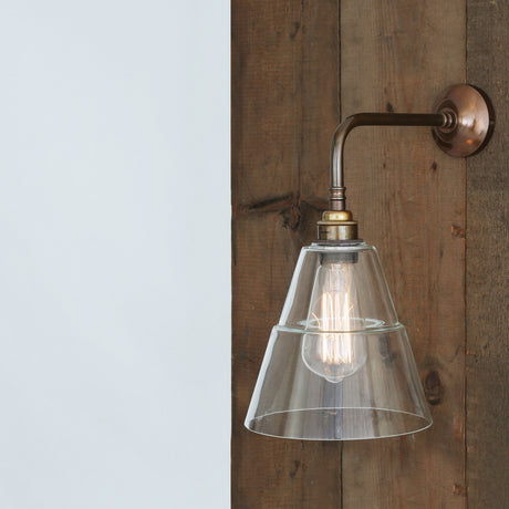Lyx Glass Wall Light with a stepped cone clear glass shade on an antique brass L-shaped arm and round wall plate, mounted on a wood-panelled wall.