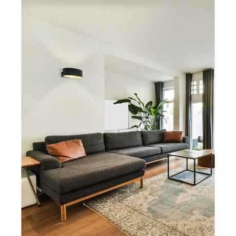 Maccacari LED wall light with an oval black outer band and gold inner finish, mounted above a dark sofa in a living room setting.
