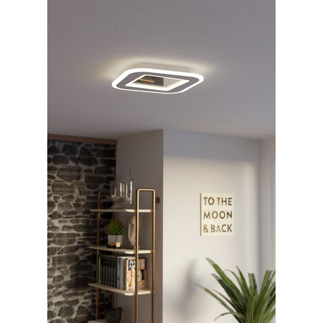 Macchioni LED Panel Light, a 38 x 38cm white square ceiling fixture with a softly glowing border, shown mounted on a plain ceiling in a living room with a bookshelf and plant below.