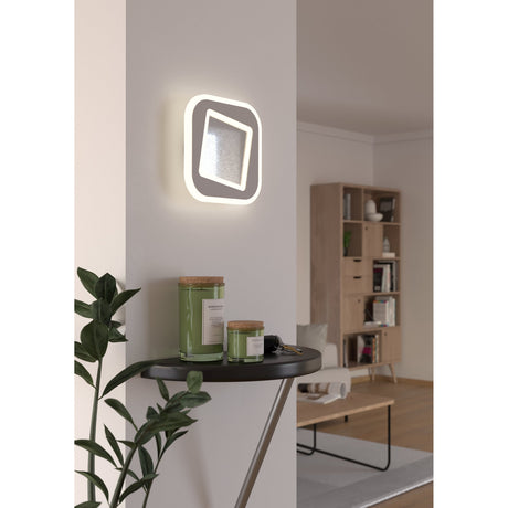 Macchioni LED Wall Light 20 x 20cm in white, a rounded square plate with a tilted inner square diffuser, shown glowing beside a console table in a living room.