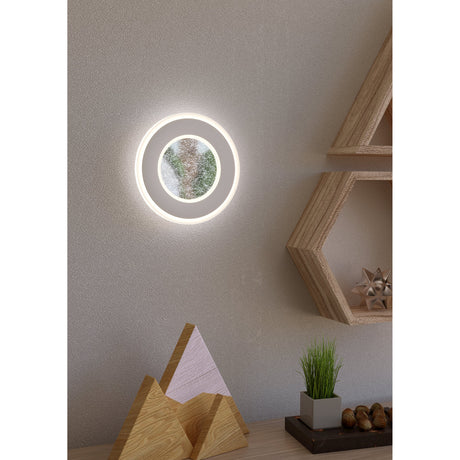 Macchioni LED Wall Light 20cm in white, round layered design with frosted outer ring and crystal-effect centre, glowing on a bedroom wall beside wooden shelving.