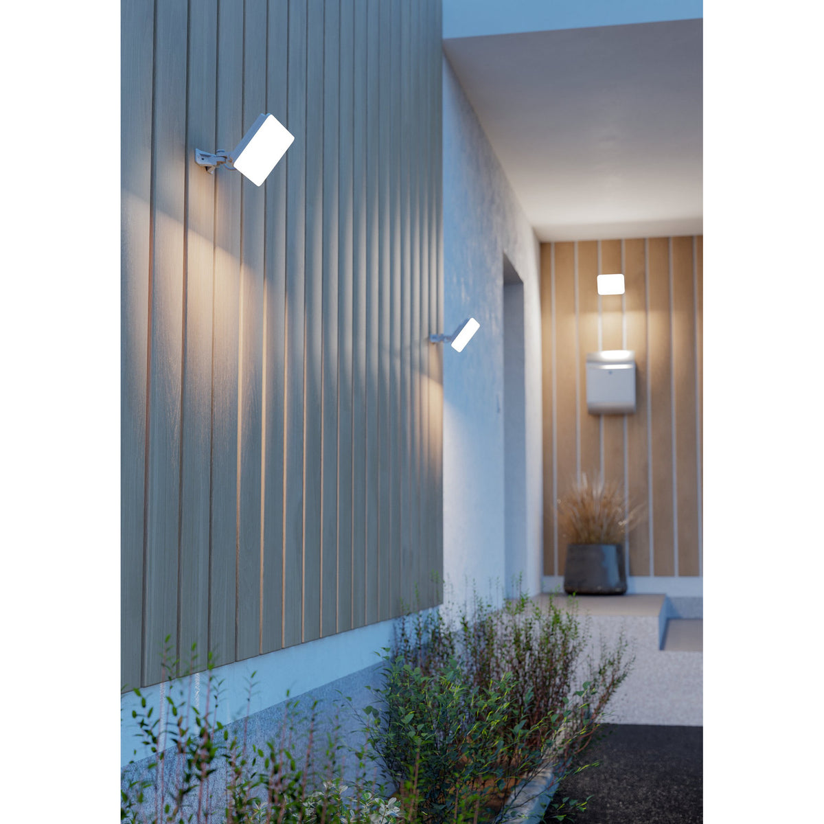 Machio LED Flood Light 30W in white, shown mounted on a slatted wall by a front entrance, with a rectangular aluminium body on an adjustable wall bracket casting warm downward light.