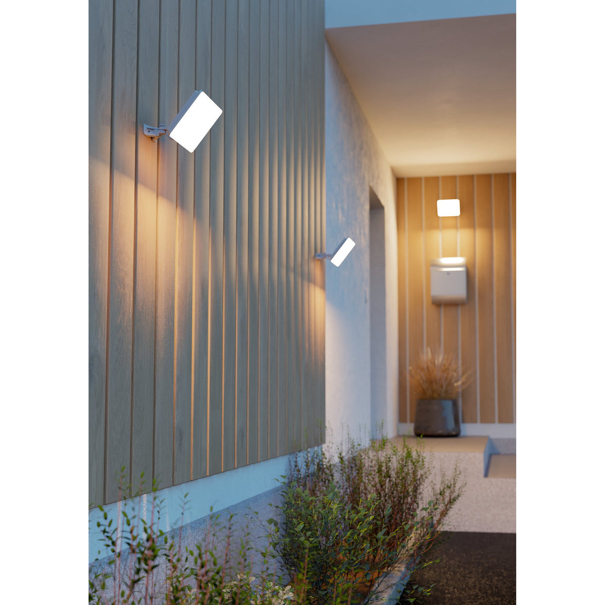 Machio 50W white LED flood light mounted on timber wall cladding near a home entrance, adjustable bracket angling the head to cast warm downward light along the path.