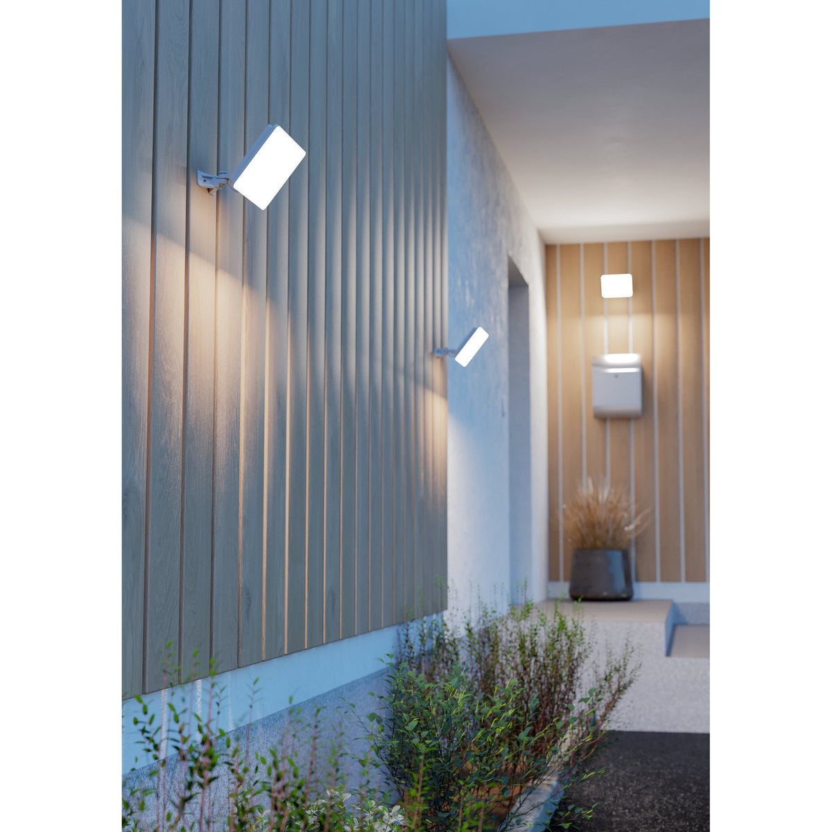 Machio LED Flood Light in white, shown mounted on adjustable wall brackets illuminating wood-slatted exterior walls along an outdoor hallway at dusk.