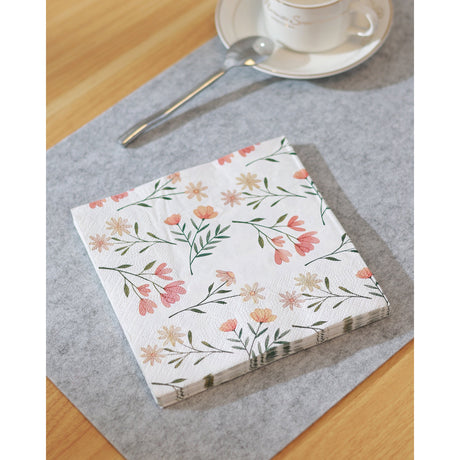 Mahuli Ditsy Floral Napkins, a folded stack of white paper napkins printed with coral and peach florals and green sprigs, set beside a teacup and spoon on a grey mat.