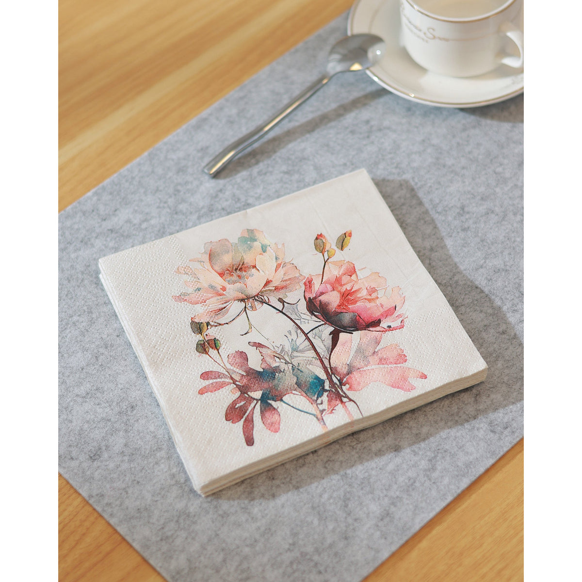 Mahuli Floral Napkins, folded square paper napkin with watercolour peony print in coral, blush and teal, shown on a grey table setting beside a spoon and cup.