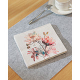 Mahuli Floral Napkins, folded square paper napkin with watercolour peony print in coral, blush and teal, shown on a grey table setting beside a spoon and cup.