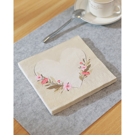 Mahuli Heart Print Napkins, folded square with a cream heart outline and pink-coral floral sprays, styled on a grey table runner beside a spoon and teacup.