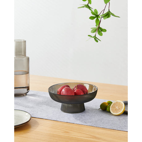 Mambrui Glass Decorative Bowl 20cm in smoky grey with ribbed pedestal base, shown holding apples on a wooden table with a runner, vases and greenery nearby.