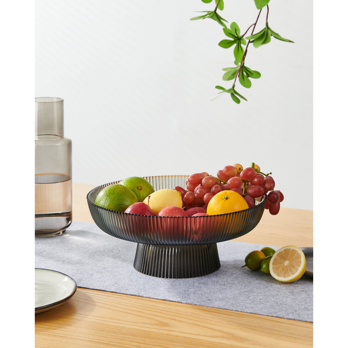 Mambrui 30cm smoky grey glass decorative bowl with ribbed footed base, filled with fruit on a wooden table with a woven runner.