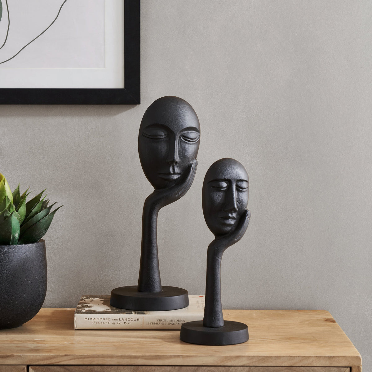 Manambato Sculpture in black aluminium, two stylised faces with closed eyes resting in cupped hands on slender stands, displayed on a wooden sideboard beside a plant and books.